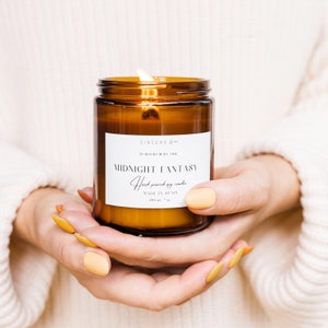 Manos femeninas sostienen una vela encendida en un frasco ámbar con etiqueta beige, vistiendo un acogedor suéter blanco