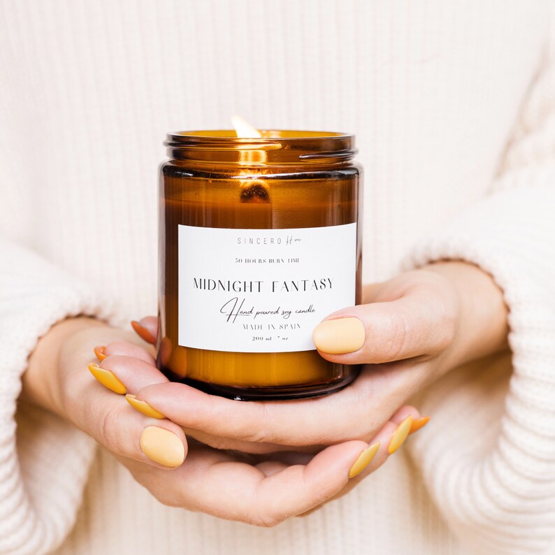 Manos femeninas sostienen una vela encendida en un frasco ámbar con etiqueta beige, vistiendo un acogedor suéter blanco