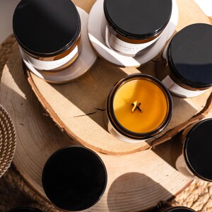 Vista superior de 9 velas en frascos ámbar, dispuestas en círculo sobre soportes de madera, con una vela encendida en el centro y sombra de ramas de fondo