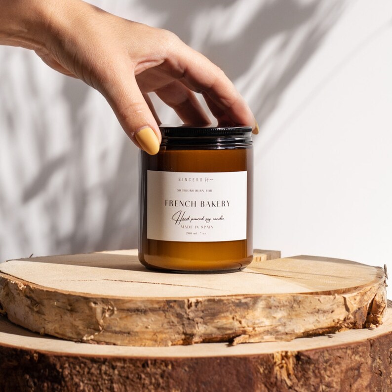 Imagen: Vela en frasco ámbar con etiqueta beige en dos soportes de madera. Mano femenina abre la elegante tapa. Fondo con sombra de ramas