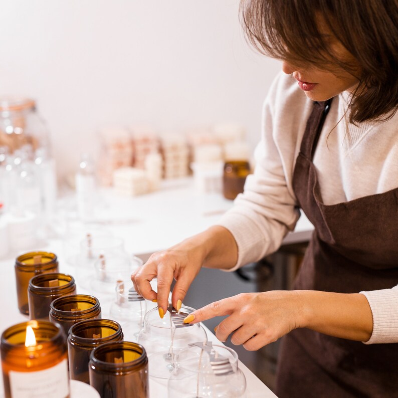 Creación de velas artesanales: mujer preparando velas en recipientes diferentes, colocando mechas en frascos. En segundo plano, productos del fabricante.