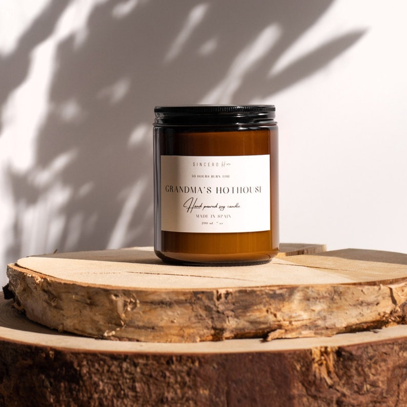 Una cautivadora imagen de una vela en un elegante frasco de ámbar con una tapa negra y una etiqueta beige exquisita. Esta lujosa vela se encuentra en dos elegantes soportes de madera, creando un fondo sofisticado con la sombra de flores pintorescas.