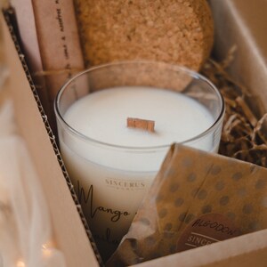Puede incluir: Una caja de cartón marrón con una vela blanca dentro. La vela está en un frasco de vidrio transparente con una mecha de madera. La caja también contiene una pequeña bolsa de papel marrón con una pegatina circular marrón que dice "BLOOM" y "SINCERUS". El frasco de la vela tiene una etiqueta que dice "SINCERUS" y "Mango & Vainilla".