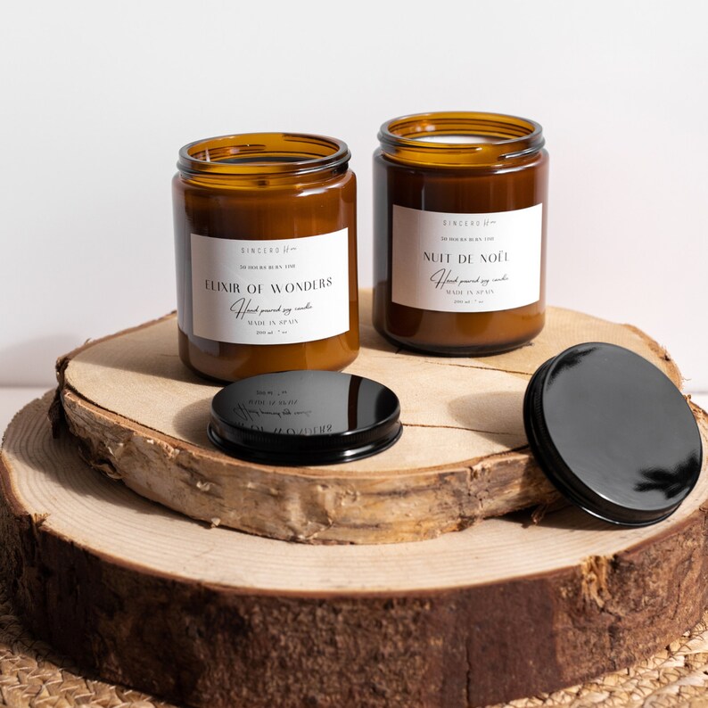 Dos lujosas velas en elegantes frascos ámbar con etiquetas beige, apoyadas sobre elegantes soportes de madera. Las tapas negras y las velas encendidas crean una atmósfera de calidez y encanto