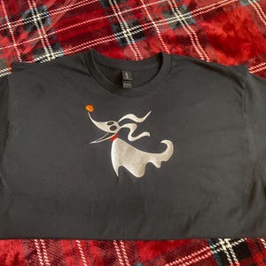 May include: Black t-shirt with a silver ghost dog design, a red collar, and an orange pumpkin detail. The graphic is centrally placed. The shirt rests on a red and black tartan blanket, a classic pattern.