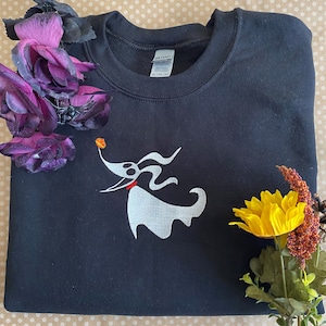 May include: Black sweatshirt featuring a white embroidered image of Zero, the ghost dog from The Nightmare Before Christmas, holding a bone. The sweatshirt has a crew neck and long sleeves.