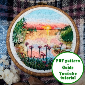 May include: Embroidered sunset landscape in a wooden embroidery hoop. The scene features a lake with reeds, a sunset sky, and pink flowers. The text "PDF pattern + Guide + Youtube tutorial" is visible in the lower right corner.