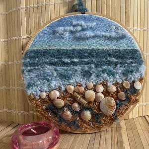 Op de afbeelding: Ronde, geborduurde wandkunst die een strandscène uitbeeldt. Het kunstwerk toont een blauwe lucht met wolken, oceaangolven en schelpen op een zandstrand. Het stuk is gevat in een houten hoepel en is een uniek woondecoratie-item.