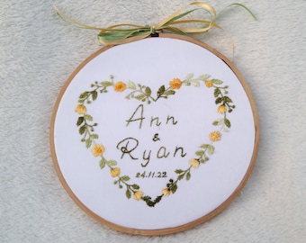 May include: Embroidered hoop with a heart-shaped wreath of yellow and green flowers. The wreath surrounds the names "Ann & Ryan" and the date "24/11/22".