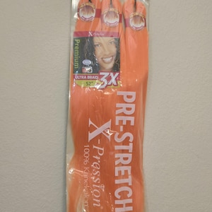 Op de afbeelding: Een verpakking Outre X-Pression voorgerekt vlecht haar in Pumpkin Spice. De verpakking toont een foto van een vrouw en de tekst "ULTRA BRAID 3X 132 cm". Het haar is fel oranje.