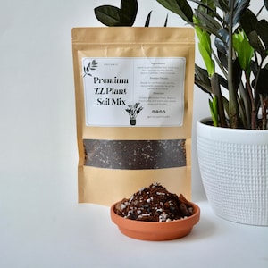 May include: A brown paper bag with a label that reads "Organic Premium ZZ Plant Soil Mix". The bag is filled with a dark brown soil mix. There is a small terracotta dish with a sample of the soil mix in front of the bag. The bag is sitting on a white surface.