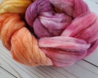 May include: A bundle of colorful wool roving in shades of yellow, orange, pink, and purple. The wool is arranged in a circular shape, with the different colors blending together.