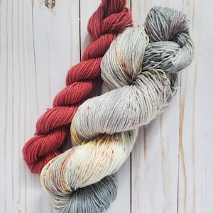 May include: Two skeins of yarn in the image. One is a solid, deep red color, and the other is a variegated yarn with shades of gray, white, yellow, and red. The yarn is arranged on a light-colored wooden surface.