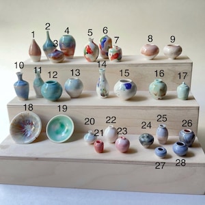 May include: A collection of small, handcrafted ceramic vases and bowls in various shapes and colors. The assortment includes miniature bottles, rounded vases, and small bowls, showcasing a range of glazes and decorative patterns. The pieces are displayed on a light wooden surface.