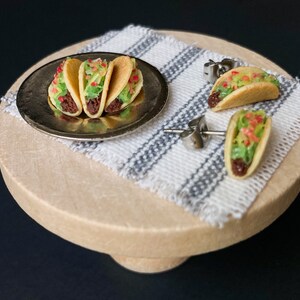 Peut inclure: Un ensemble de trois boucles d'oreilles miniatures en forme de tacos. Les boucles d'oreilles sont en p&acirc;te polym&egrave;re et pr&eacute;sentent un design r&eacute;aliste de taco avec une garniture brune, de la laitue verte et une tomate rouge. Les boucles d'oreilles sont pr&eacute;sent&eacute;es sur un pr&eacute;sentoir en bois avec un tissu ray&eacute; blanc et gris.