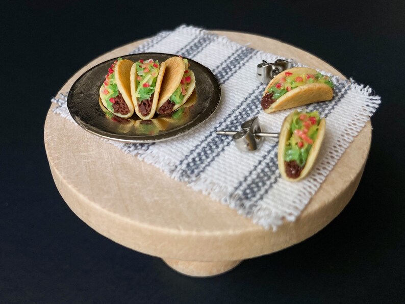 Peut inclure: Un ensemble de trois boucles d'oreilles miniatures en forme de tacos. Les boucles d'oreilles sont en p&acirc;te polym&egrave;re et pr&eacute;sentent un design r&eacute;aliste de taco avec une garniture brune, de la laitue verte et une tomate rouge. Les boucles d'oreilles sont pr&eacute;sent&eacute;es sur un pr&eacute;sentoir en bois avec un tissu ray&eacute; blanc et gris.