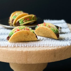 Peut inclure: Deux tacos miniatures garnis de viande brune, de laitue verte et de tomates rouges. Les tacos sont sur une surface ray&eacute;e blanche et grise.
