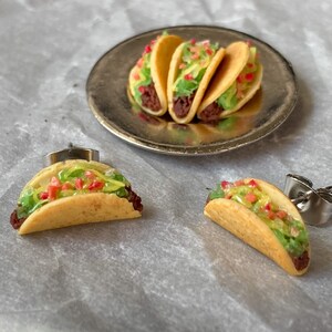 Peut inclure: Une paire de boucles d'oreilles repr&eacute;sentant des tacos miniatures avec de la viande brune, de la laitue verte et des garnitures de tomates rouges. Les tacos sont faits de p&acirc;te jaune et sont pr&eacute;sent&eacute;s sur une assiette en argent.