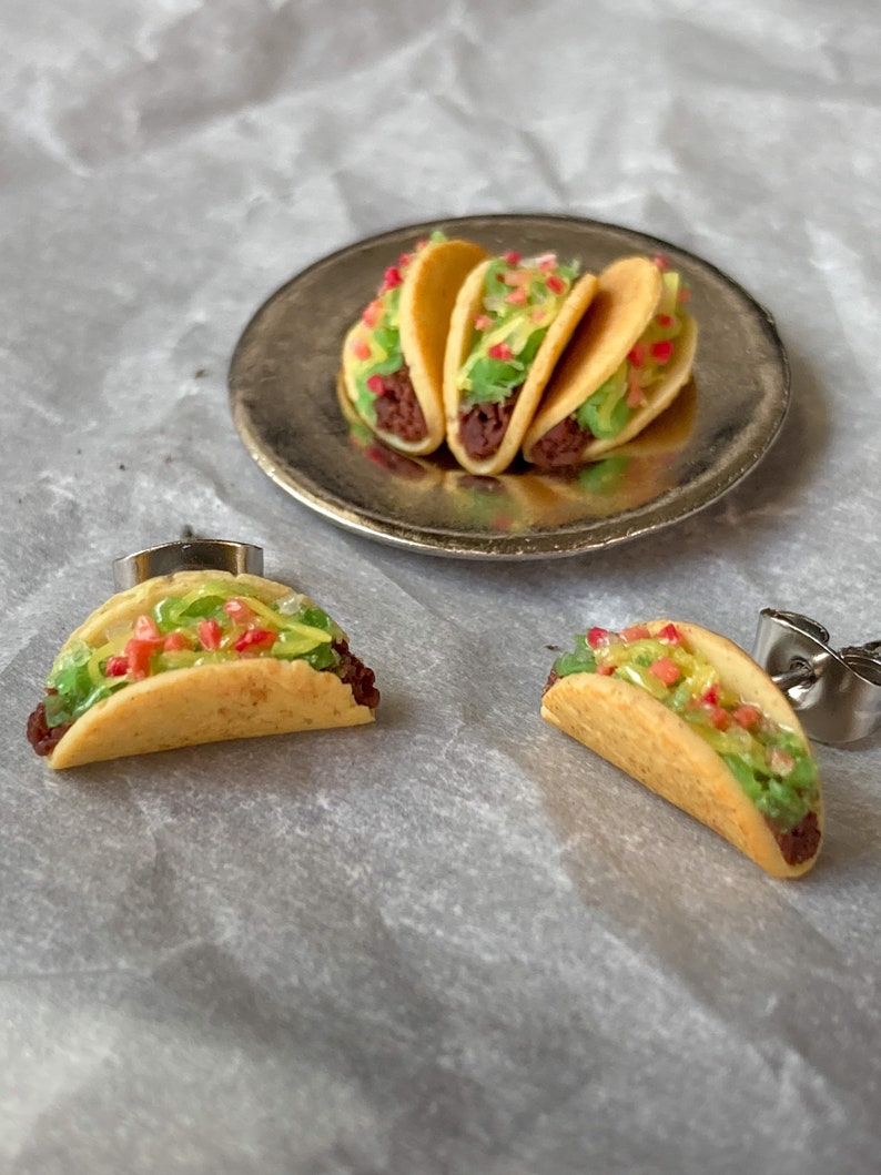 Peut inclure: Une paire de boucles d'oreilles repr&eacute;sentant des tacos miniatures avec de la viande brune, de la laitue verte et des garnitures de tomates rouges. Les tacos sont faits de p&acirc;te jaune et sont pr&eacute;sent&eacute;s sur une assiette en argent.