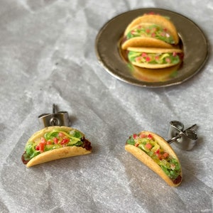 Peut inclure: Une paire de boucles d'oreilles en forme de taco argent&eacute;es avec de la laitue verte, des tomates rouges et du fromage jaune. Les boucles d'oreilles sont sur un fond blanc.