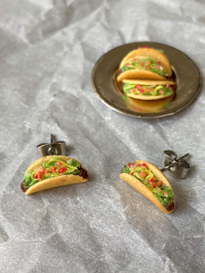 Peut inclure: Une paire de boucles d'oreilles en forme de taco argent&eacute;es avec de la laitue verte, des tomates rouges et du fromage jaune. Les boucles d'oreilles sont sur un fond blanc.