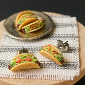 Peut inclure: Une paire de boucles d'oreilles en forme de tacos miniatures avec des tiges argent&eacute;es. Les tacos sont en p&acirc;te polym&egrave;re et pr&eacute;sentent une coque brune, de la laitue verte, une tomate rouge et du fromage jaune.