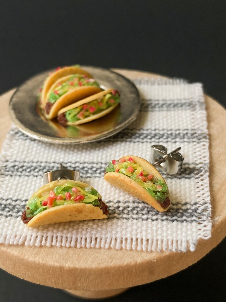 Peut inclure: Une paire de boucles d'oreilles en forme de tacos miniatures avec des tiges argent&eacute;es. Les tacos sont en p&acirc;te polym&egrave;re et pr&eacute;sentent une coque brune, de la laitue verte, une tomate rouge et du fromage jaune.