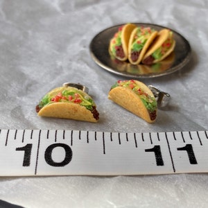 Peut inclure: Une paire de boucles d'oreilles en forme de taco miniature, chacune avec une coquille de taco brune, jaune et verte remplie de garnitures rouges et vertes. Les boucles d'oreilles sont pr&eacute;sent&eacute;es &agrave; c&ocirc;t&eacute; d'une r&egrave;gle, les boucles d'oreilles en forme de taco mesurant environ 2,5 cm de long.