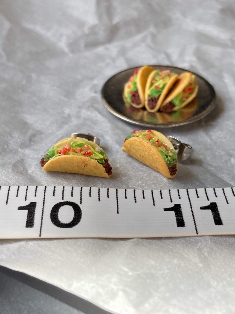 Peut inclure: Une paire de boucles d'oreilles en forme de taco miniature, chacune avec une coquille de taco brune, jaune et verte remplie de garnitures rouges et vertes. Les boucles d'oreilles sont pr&eacute;sent&eacute;es &agrave; c&ocirc;t&eacute; d'une r&egrave;gle, les boucles d'oreilles en forme de taco mesurant environ 2,5 cm de long.