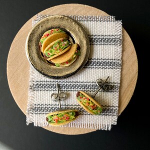 Peut inclure: Une paire de boucles d'oreilles en forme de tacos avec une assiette de tacos en arri&egrave;re-plan. Les boucles d'oreilles sont en p&acirc;te polym&egrave;re et sont peintes pour ressembler &agrave; de vrais tacos. Les tacos sont garnis de viande, de laitue et de tomate. Les boucles d'oreilles sont sur un tissu ray&eacute; blanc et gris.