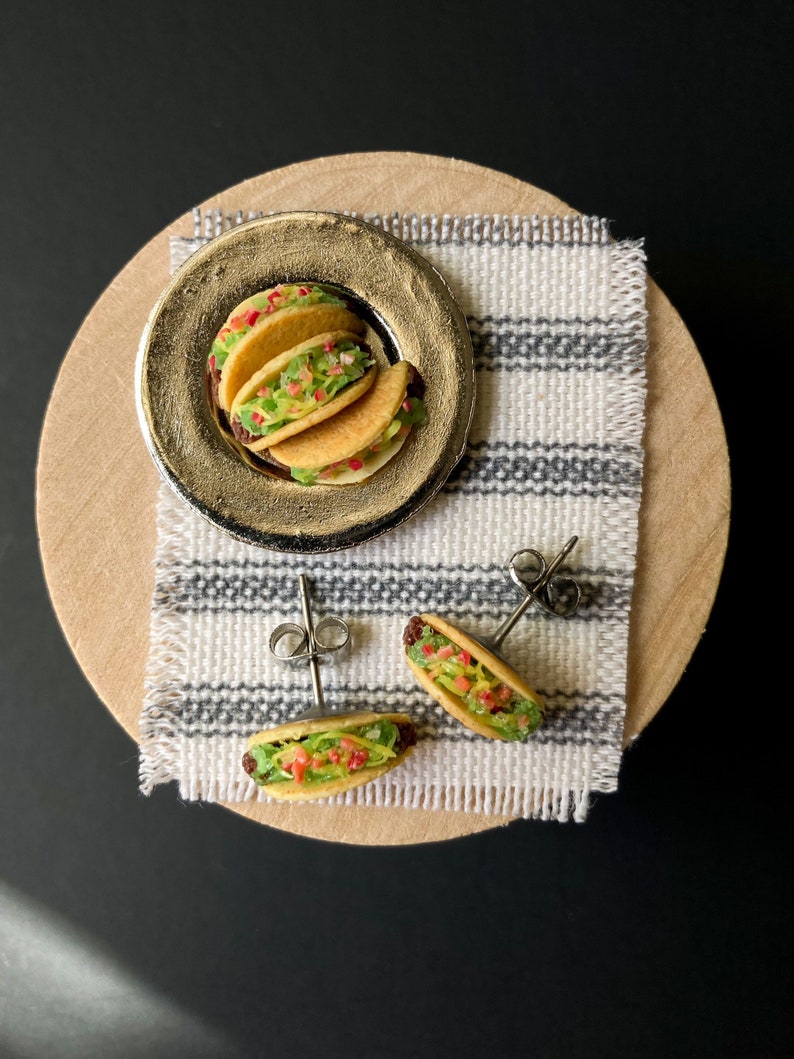Peut inclure: Une paire de boucles d'oreilles en forme de tacos avec une assiette de tacos en arri&egrave;re-plan. Les boucles d'oreilles sont en p&acirc;te polym&egrave;re et sont peintes pour ressembler &agrave; de vrais tacos. Les tacos sont garnis de viande, de laitue et de tomate. Les boucles d'oreilles sont sur un tissu ray&eacute; blanc et gris.