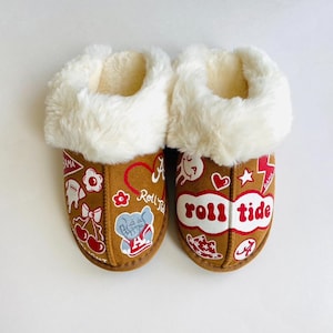 May include: A pair of brown suede slippers with white faux fur trim. The slippers are decorated with various graphics and the words "Roll Tide" in red and white. The interior is lined with a soft, cream-colored material.