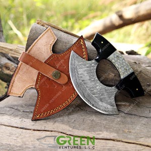 May include: A Damascus steel axe with a black and white handle and a leather sheath. The axe has a unique, wavy pattern on the blade.