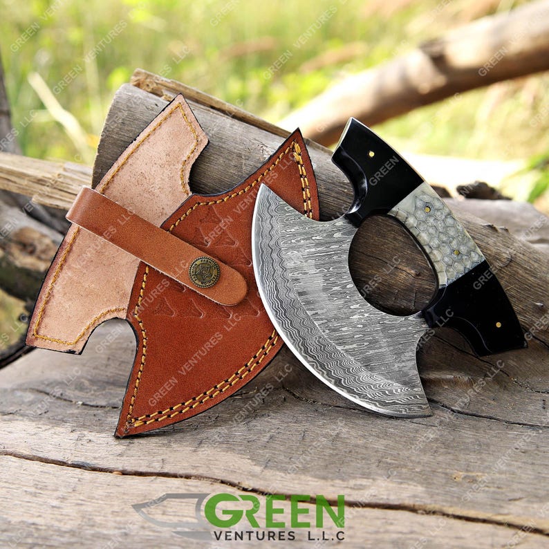 May include: A Damascus steel axe with a black and white handle and a leather sheath. The axe has a unique, wavy pattern on the blade.