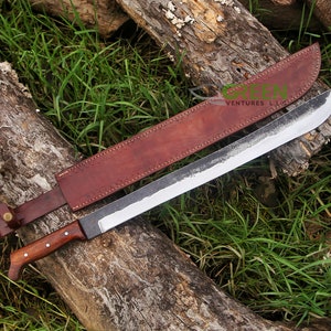 May include: A large, hand-forged machete with a dark, textured blade and a wooden handle. It rests on a log with a brown leather sheath. The blade is approximately 61 cm long.