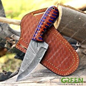 May include: A small, fixed blade knife with a Damascus steel blade and a purple and orange wood handle. The knife is in a brown leather sheath.