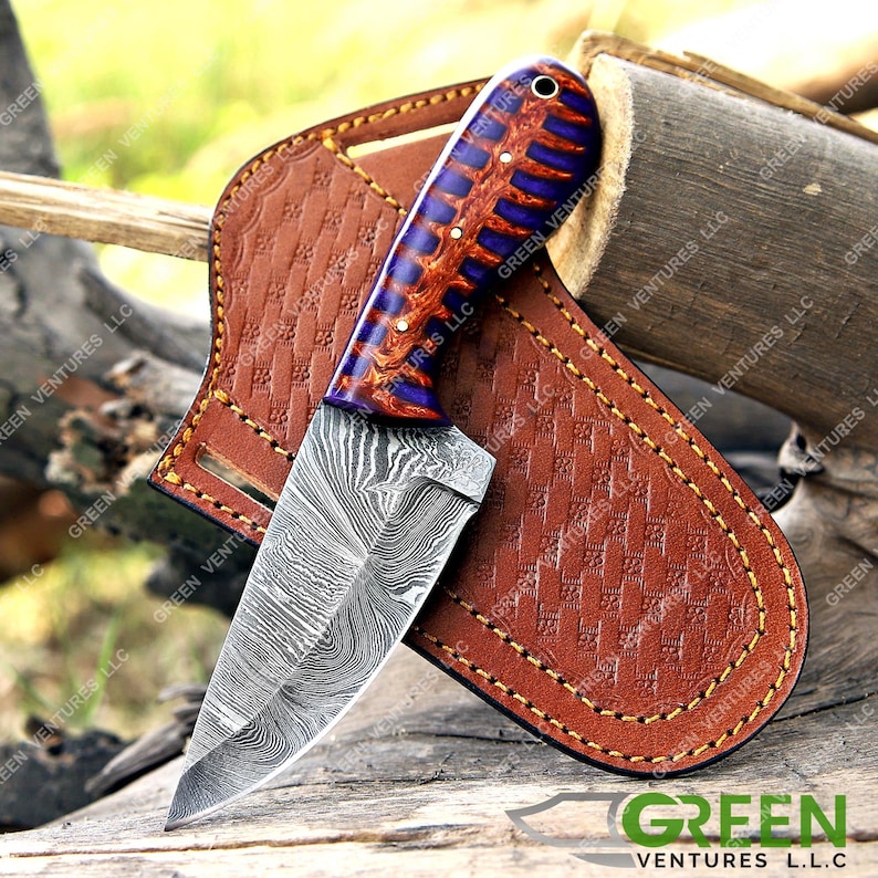 May include: A small, fixed blade knife with a Damascus steel blade and a purple and orange wood handle. The knife is in a brown leather sheath.