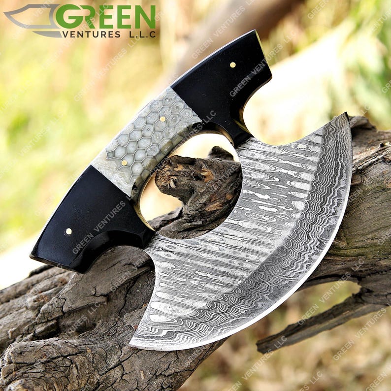May include: A black and white handled cleaver with a Damascus steel blade. The handle has a honeycomb pattern and the blade has a wavy pattern. The cleaver is resting on a piece of wood.