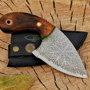 May include: A small, curved knife with a brown wooden handle and a silver blade. The blade has a decorative pattern of snowflakes and is partially covered by a black leather sheath. The sheath has a brass button and a brass tag with the word "FASHION" on it.