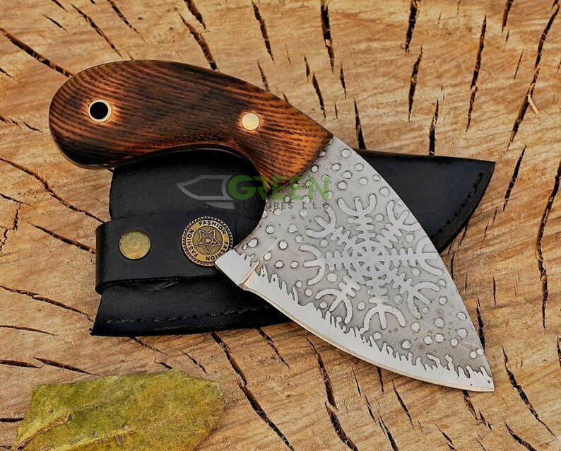 May include: A small, curved knife with a brown wooden handle and a silver blade. The blade has a decorative pattern of snowflakes and is partially covered by a black leather sheath. The sheath has a brass button and a brass tag with the word "FASHION" on it.