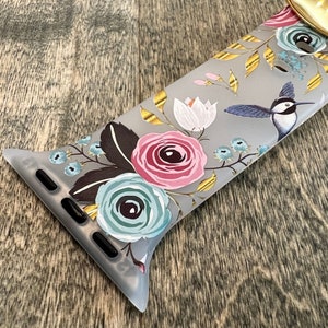 May include: A gray Apple Watch band with a floral print featuring pink, blue, and white flowers, gold accents, and a hummingbird.