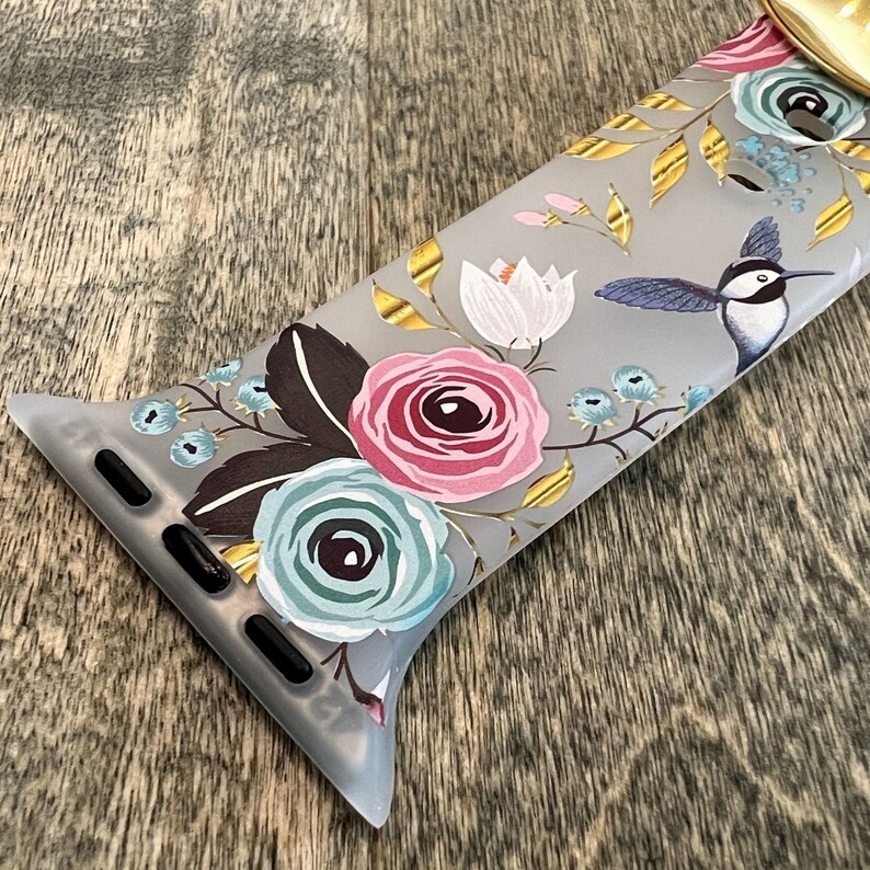 May include: A gray Apple Watch band with a floral print featuring pink, blue, and white flowers, gold accents, and a hummingbird.