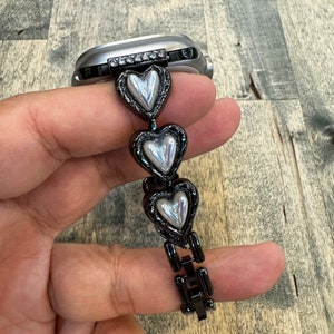 Könnte beinhalten: Schwarzes und silbernes Armband in Herzform mit einem silbernen und schwarzen Verschluss. Das Armband ist aus Metall und hat eine glänzende Oberfläche.