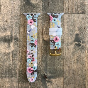 May include: A floral patterned Apple Watch band with a gold buckle. The band is made of silicone and has a light gray background with pink, blue, and green flowers.