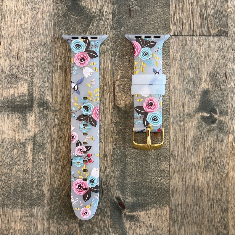 May include: A floral patterned Apple Watch band with a gold buckle. The band is made of silicone and has a light gray background with pink, blue, and green flowers.