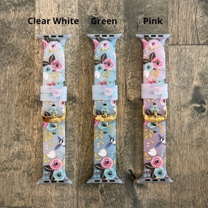 May include: Three Apple Watch bands with a floral pattern. The bands are in clear white, green, and pink. The bands have a gold buckle.