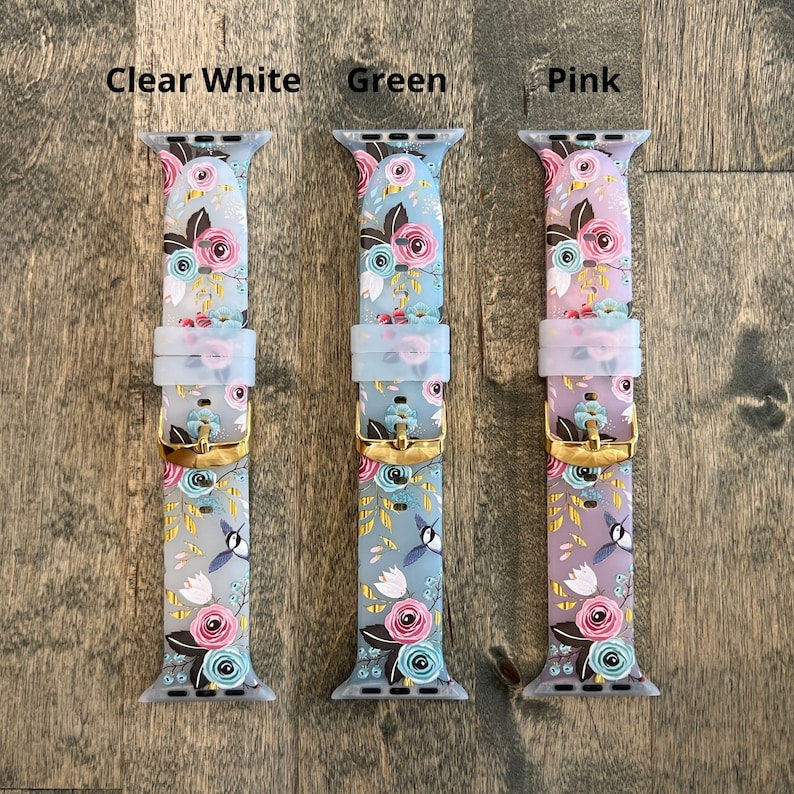May include: Three Apple Watch bands with a floral pattern. The bands are in clear white, green, and pink. The bands have a gold buckle.
