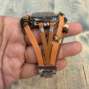 Puede incluir: Una correa de reloj de cuero marrón y beige con una esfera plateada. La correa presenta múltiples correas de cuero, cuentas de madera y un dije de estrella dorada. La correa del reloj se sostiene en una mano.