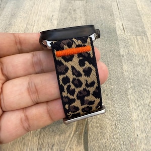 May include: A black smartwatch with a leopard print strap. The strap has a black background with tan and brown spots, and an orange stripe. The watch has a black case and silver hardware.