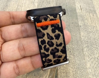 Peut inclure: Une montre intelligente noire avec un bracelet à imprimé léopard. Le bracelet présente un fond noir avec des taches beiges et marron, et une bande orange. La montre a un boîtier noir et des éléments argentés.